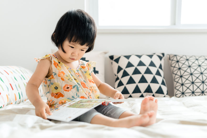 Cute Toddler Girl Reading Book On Bed